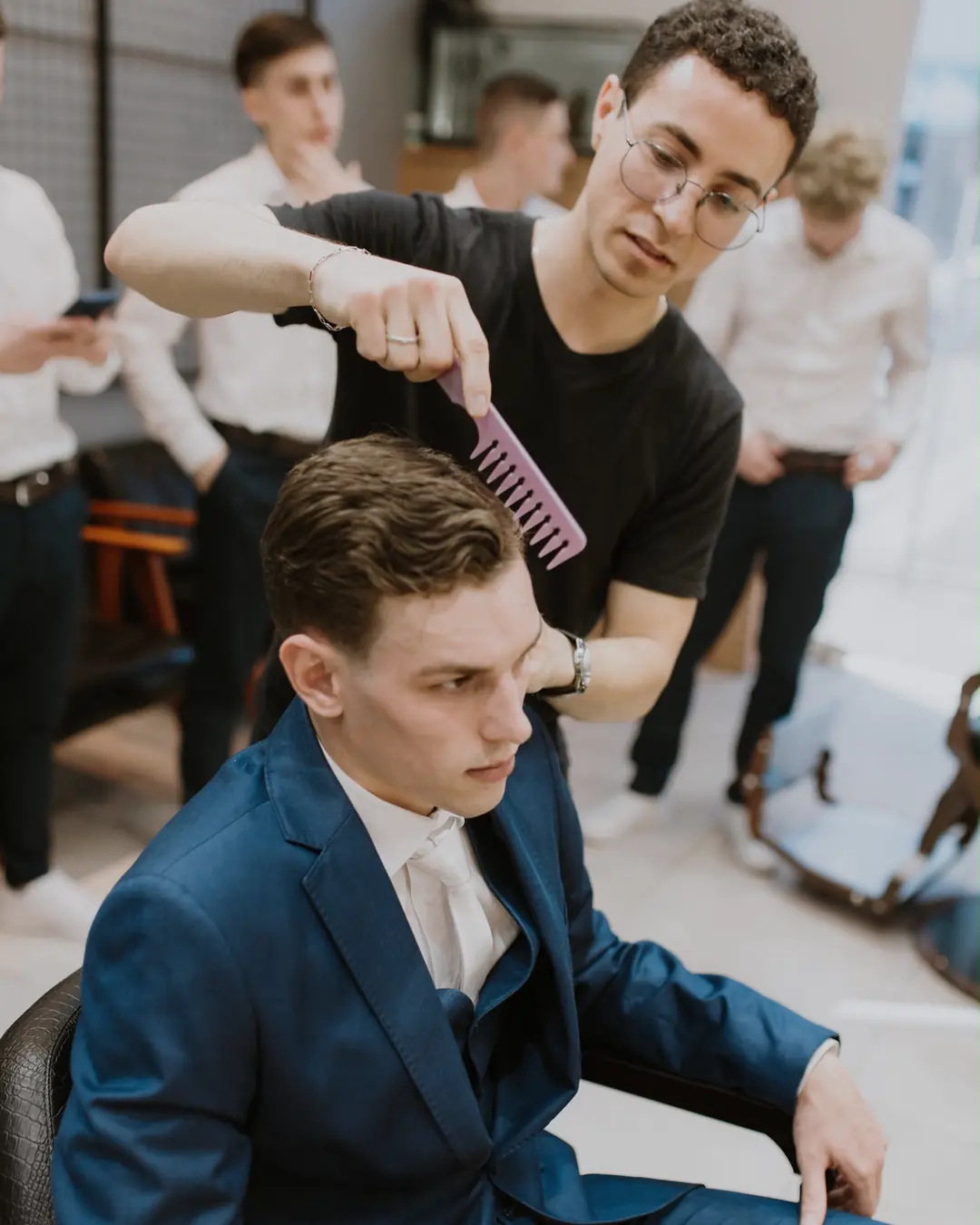 João finalizando o penteado de um noivo de terno azul, com os padrinhos ao fundo
